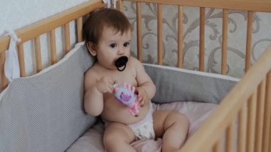 Ciddi bir bebeğin portresi. Beşiğinde bir oyuncakla oturmuş emzik emiyor. Bebek bakımı. Sağlıklı yemek..