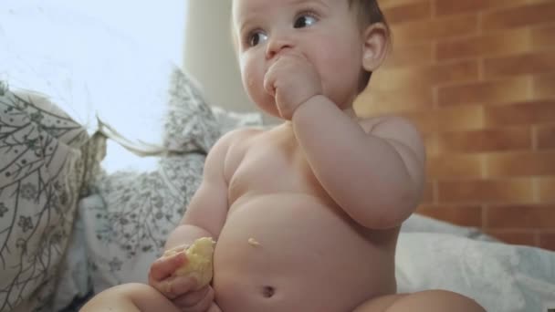 Portret van een klein meisje geniet van het eten van een banaan met een zoete glimlach zittend binnen op een bed. Vers gezond voedsel. Baby ontwikkeling. Gezond dieet. Leuk klein meisje. — Stockvideo