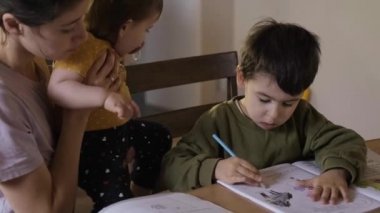 Anaokulu çocuğu masada oturuyor, albümde resim çiziyor, beyaz bir anne ve küçük bir kız çocuğu çocuğun yanında oturuyor. Güzel genç kız.