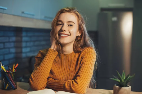 Donna dai capelli rossi seduta a tavola con taccuino e guardando la fotocamera sognante. Riflettere sulle idee creative per la ricerca. — Foto Stock