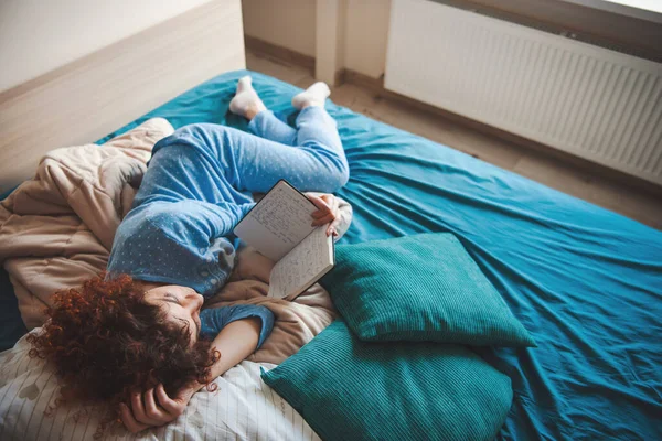 Mujer caucásica de pelo rizado acostada cómodamente en la cama leyendo un libro. Libro de lectura de mujer rizada para diseño de estilo de vida de moda. — Foto de Stock