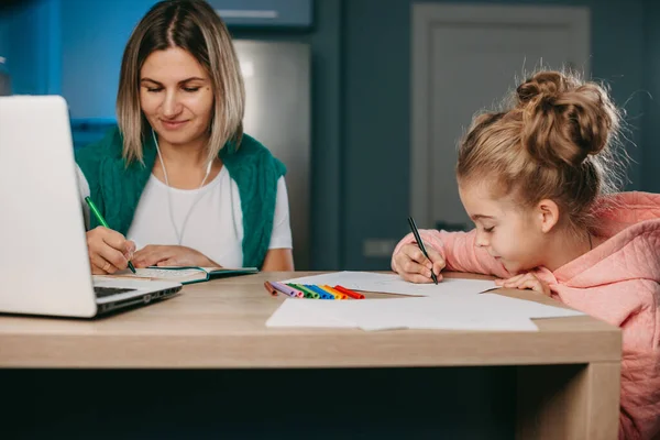 Mujer con auriculares trabajando remotamente en el portátil mientras su hija dibuja con lápices de colores a su lado. freelance, trabajo online, estudio, aprendizaje con —  Fotos de Stock