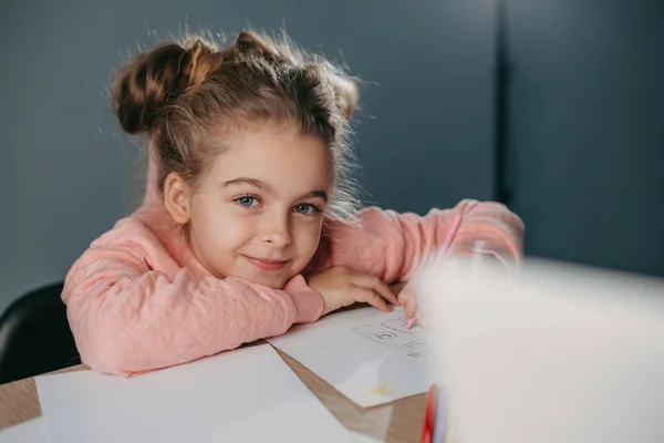 Retrato de una colegiala sentada a la mesa y dibujando con lápices de colores en casa mirando a la cámara y sonriendo. Una idea creativa. Sonriente niña feliz. Disfrutando —  Fotos de Stock