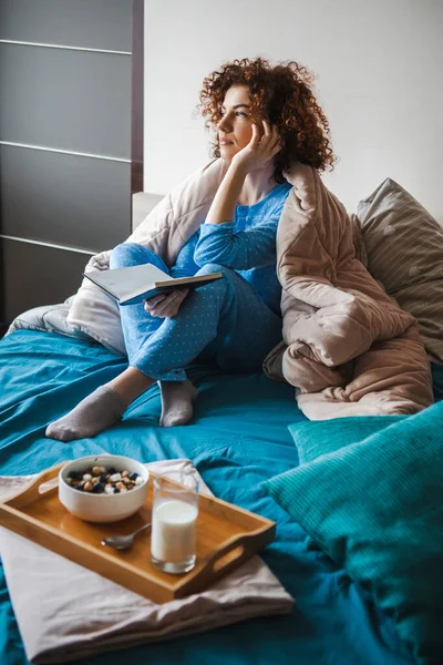 Σγουρομάλλα μελαχρινή γυναίκα κάθεται στο κρεβάτι το πρωί με το βιβλίο στο χέρι, με πρωινό στο κρεβάτι. Για σχεδιασμό τρόπου ζωής. Υγιεινή τροφή. Βιολογικά τρόφιμα. — Φωτογραφία Αρχείου