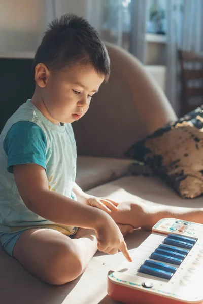 Garoto caucasiano brincando com seu teclado favorito piano brinquedo sentado no sofá em casa. Brinquedos educativos para conceito de crianças. Foco seletivo. — Fotografia de Stock