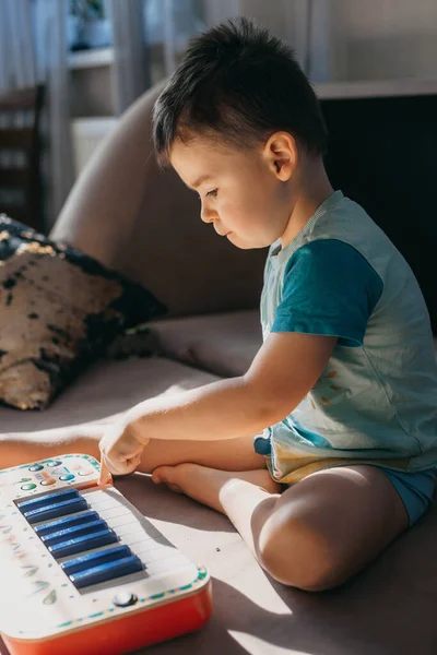 Menino caucasiano sentado em uma cama e brincando com um piano de música de brinquedo de plástico. Conceito de educação. Foco seletivo. — Fotografia de Stock