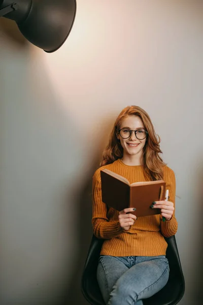 Zrzavá žena sedící na židli nad bílou stěnou a čtoucí knihu pózující ve světle reflektorů. Obchod, vzdělávací koncept. Moderní kreativní podnikatelský koncept — Stock fotografie