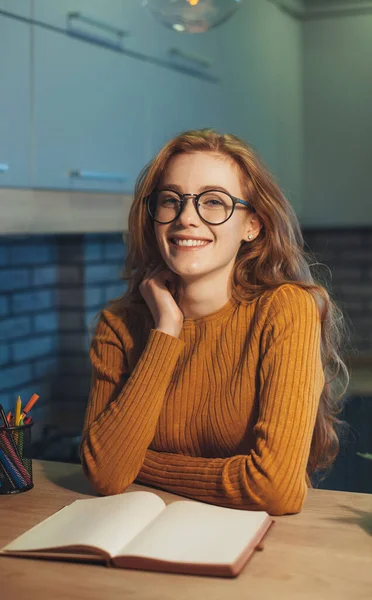 Šťastná studentka, která sedí u stolu a usmívá se na kameru, zatímco čte papírovou knížku. Krásná tvářička. Obchodní portrét. — Stock fotografie