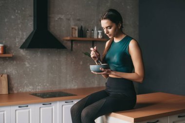 Sportif bir kadın mutfakta elinde bir kase ve kaşıkla yemek yiyor. Vejetaryen sağlıklı yiyecekler. Sağlıklı bir yaşam tarzı. Sağlıklı beslenme, diyet, kilo.