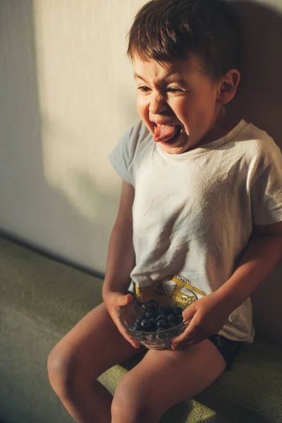 Boy eating blueberries on the couch and grimacing that he eat a sour blueberry. Facial expression. — стоковое фото