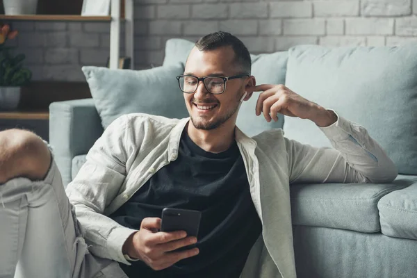 Hombre feliz usando anteojos usando el teléfono celular mientras se relaja en el suelo en casa, escuchando musicaciones en auriculares inalámbricos. Uso de auriculares y teléfono móvil —  Fotos de Stock