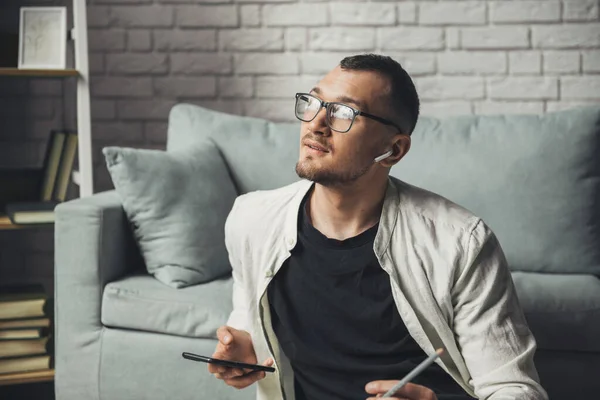 Homem caucasiano de óculos usando telefone celular enquanto trabalhava sentado no chão ouvindo música com fones de ouvido sem fio. — Fotografia de Stock