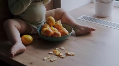 Bebek kız, bacaklarının arasında bir kâseyle portakal kabuğunun yanında oturmuş mandalina yiyor. Taze sağlıklı yiyecekler. Organik yiyecekler. Tatlı yemek..