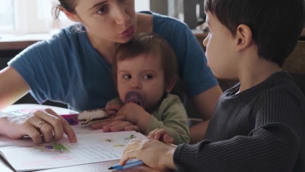 Küçük çocuk annesinin küçük yardımıyla matematik ödevini yapıyor. Anne oğluna matematik problemini açıklarken, aynı zamanda bebeği için de endişeleniyor. — Stok video