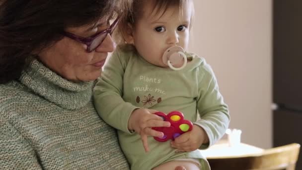 Dziewczynka ssąca smoczek i bawiąca się wielobarwną zabawką trzymaną przez babcię w okularach. Opieka nad dziećmi. Zdrowy styl życia. Portret z widokiem z boku. — Wideo stockowe