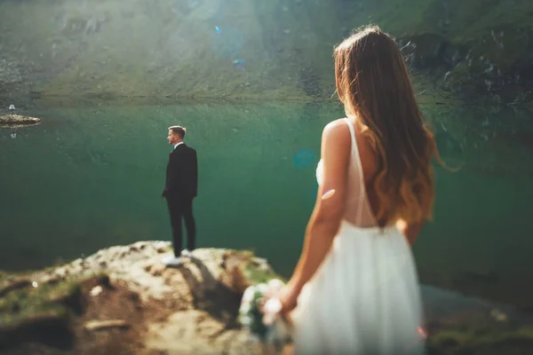 Η νύφη πλησιάζει πίσω από τον γαμπρό, ο οποίος περιμένει στην πλαγιά του βουνού κοιτάζοντας το τοπίο με τα βουνά και τη λίμνη. Φυσικό τοπίο. Γάμος — Φωτογραφία Αρχείου