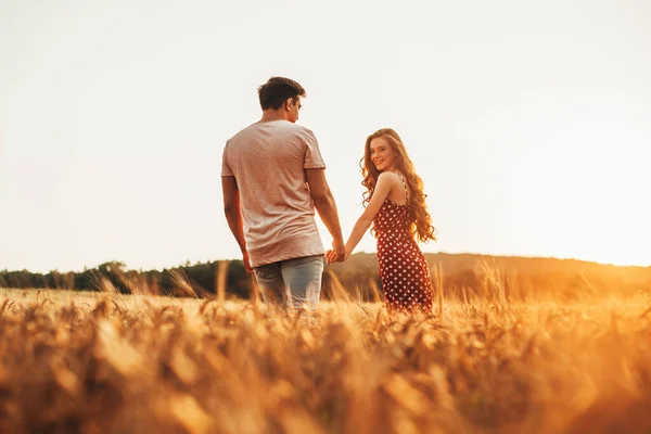 Happy woman and man walking in wheat ears field in sunny day holding hands enjoying romantic moment. Portrait of smiling red-headed woman. Nature landscape — Fotografia de Stock