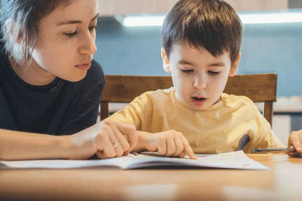 Caucasian little boy and his mother sitting together at table and reading book, mother teaching her child to read. Education, learning, study. Mother language. — Fotografia de Stock