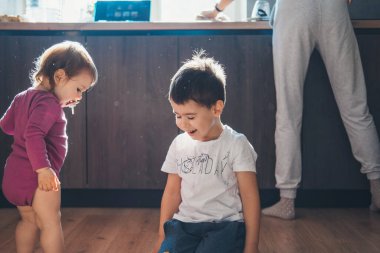 Portrait of a boy with his baby sister sitting in the kitchen and having fun with each other while mom prepares breakfast. Family care. Baby care.