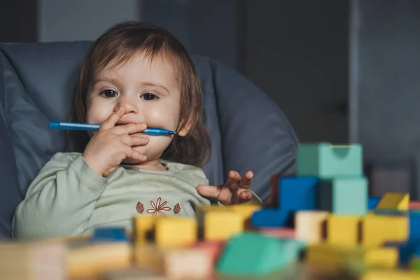 Bebê menina segurando uma caneta em sua boca, enquanto brinquedos de diferentes formas de construção sentar em sua mesa. Desenvolvimento do bebé. Retrato tirado. — Fotografia de Stock