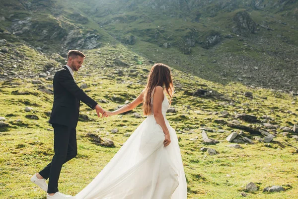 Long-haired bride in wedding dress and groom holding hands and walking in mountainside at sunset. Happy wedding couple — Zdjęcie stockowe