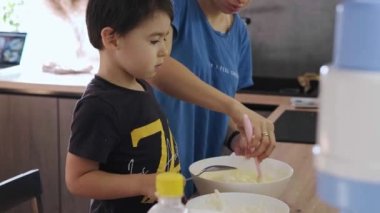 Mother with her son in their kitchen making cookies, holding a mixing bowl adding ingredients. Caucasian boy helping mother.