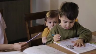 Ders masasında çizim yapan bir çocuk, küçük kız kardeşini alnından öpüyor ve arkasında oturuyor. Renkli bir portre. Bebek gelişimi. El çizimi.