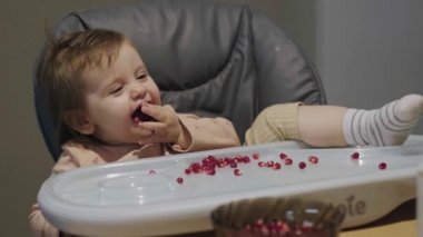 Red pomegranate seeds are thrown on the table of the baby, who eats with appetite. Baby girl sitting in a chair laughing and putting her index finger to her