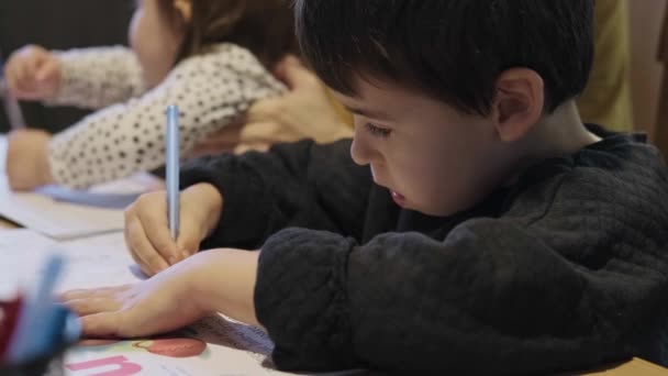 Det är dags för läxor. Grundskolepojken gör sina läxor med sin mamma, som också tar hand om sin barnsyster. Skolpojke skriva sitter på — Stockvideo