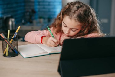 Liseli kız bilgisayar tabletinin önünde kalemle not yazıyor. Çocuk çevrimiçi öğrenim okulu öğreniyor. Uzaklık eğitimi.