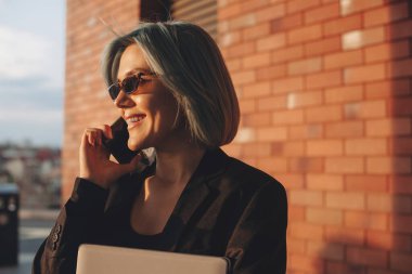Mavi saçlı, gülümseyen ve dışarıdaki tuğla duvarda duran akıllı telefonda konuşan bir kadın. Cep telefonu. Mobil iletişim. Konuşan insanlar. İş kadını.