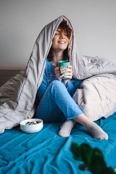 Curly-haired woman is holding a cup and smiling covering with a warm blanket. Morning coffee.