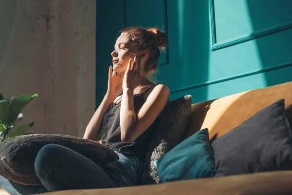 Red-haired woman applying facial paper mask sitting on the couch at home. Healthy hair. Facial skincare. — Foto de Stock