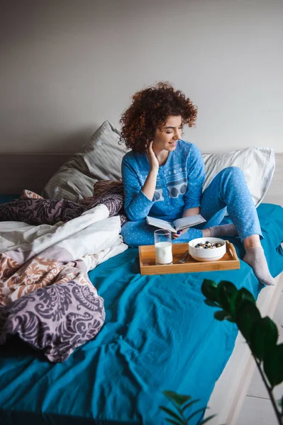 Mujer sentada en la cama desayunando con un vaso de leche y un tazón de gachas. Mañana en un acogedor y cálido dormitorio leyendo un libro. Hora de dormir. — Foto de Stock