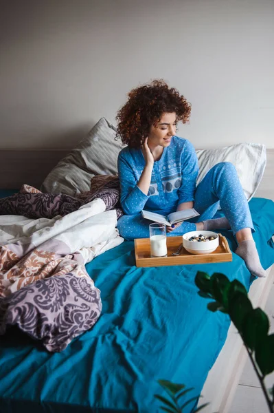 Woman sitting on bed having breakfast with glass of milk and bowl of porridge. Morning in a cozy warm bedroom reading a book. Bedtime. — Foto de Stock