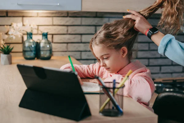 带着平板电脑的女孩在笔记本上写字，而她的母亲在整理她的头发。远距离教育。教育、学习、学习。孩子在网上学习 — 图库照片