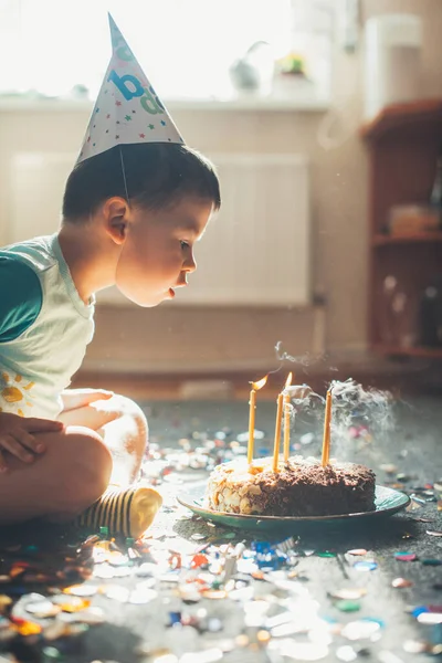 Side view of a boy celebrating his birthday, blowing candles on cake, sitting on the floor indoor. Birthday party. Fire flame. — Stock Photo, Image