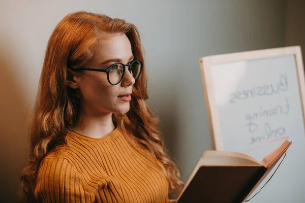 Femme aux cheveux roux assise près d'un tableau blanc et tenant un livre dans sa main lisant le nouveau sujet aux étudiants. Portrait de femme d'affaires. Éducation, apprentissage — Photo