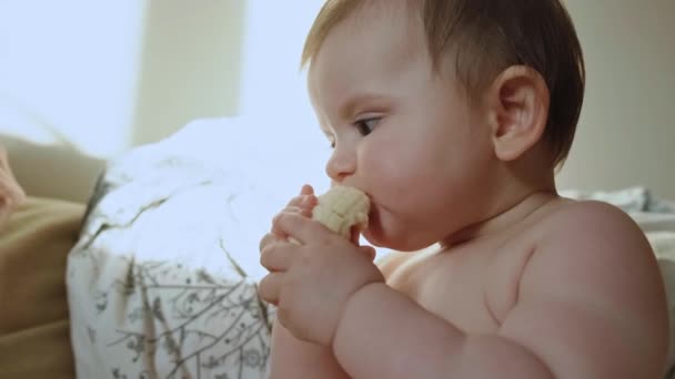 Hungrig bebis som sitter på golvet och äter en bit banan. Närbild porträtt. Vegetariskt hälsosam mat. — Stockvideo