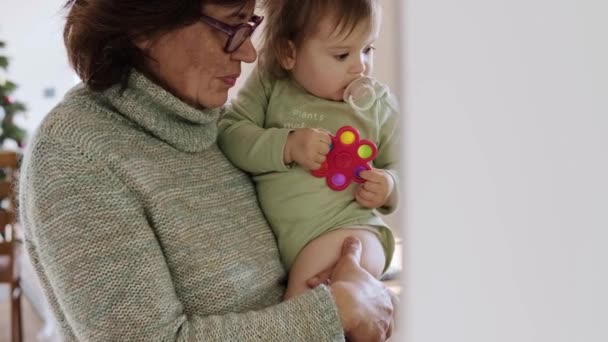 Vista de um lado de uma avó segurando uma menina em seus braços com um brinquedo. Idosos de pé juntos. Conceito familiar. — Vídeo de Stock