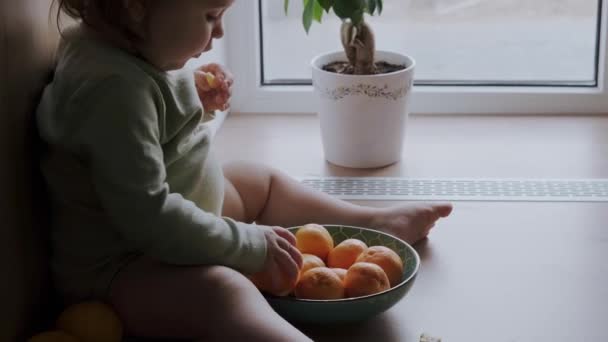 Θέα από τη μία πλευρά ενός μικρού κοριτσιού που κάθεται στο πάτωμα του δωματίου με ένα μπολ γεμάτο πορτοκάλια και τρώει με κέφι από αυτά. Φροντίδα μωρών. — Αρχείο Βίντεο