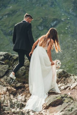 Dağdaki kayalıklarda yürüyen yeni evliler zirveye ulaşmak ve doğayı yukarıdan görmek için. Düğün seyahati. Manzara fotoğrafçılığı. Doğa manzarası