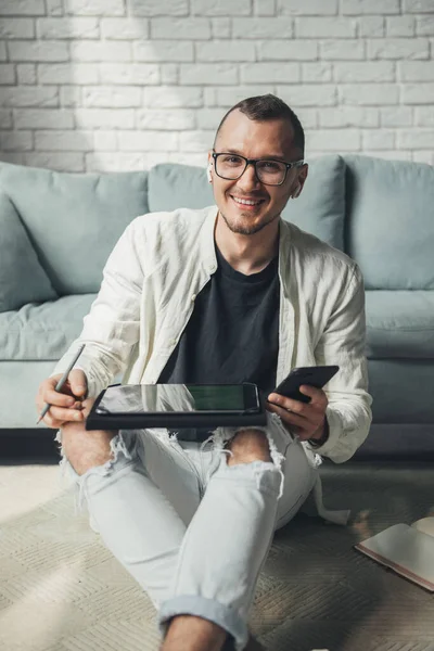 Homem olhando e sorrindo para a câmera, segurando smartphone, trabalhando em tablet sentado no chão. Conceito de marketing. Homem de negócios a trabalhar. Estilo de vida moderno. Moderno — Fotografia de Stock