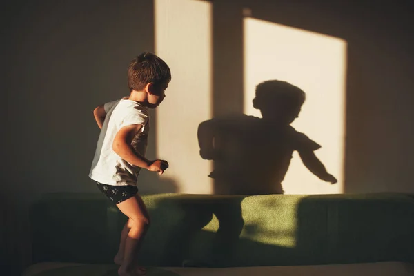 Boy playing on the couch, jumping and looking at his shadow on the wall. Sunny home. Children education. —  Fotos de Stock