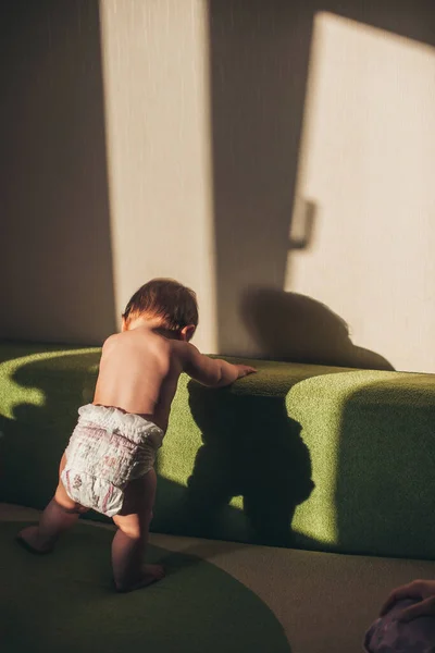 Back view baby girl standing on a sofa in the living room only in a diaper. Happy family leisure. Child development. Family care. Baby development. — Photo