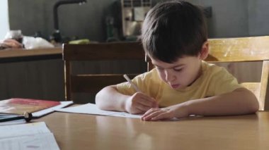 Caucasian boy using a pencil to write in the notebook sitting at the desk in the kitchen room. Front view. Coloring book.