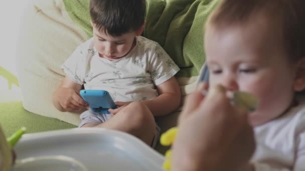 Son playing a cell phone at the same time woman feeding her baby daughter. Family concept. Motherhood concept. Baby care. Parenthood concept. Body care. — Vídeo de stock