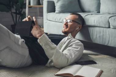 Kablosuz kulaklıklı bir adam yerde yatarken cihazlar kullanıyor, konuşuyor ve gülümsüyor. İnternet teknolojisi. İş Portresi.