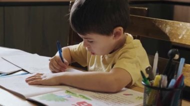 Beyaz bir çocuğun portresi alfabe yazma pratiği yapıyor evde, masada oturuyor. Mesafe çalışması. Okuma, yazma ya da çizim, evde eğitim.