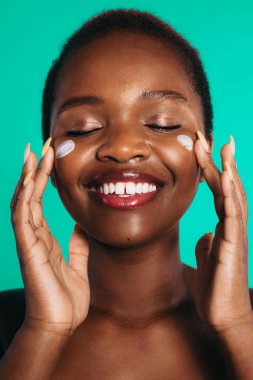 Yüzüne krem sürerek poz veren Afro-Amerikan kadının yakın plan portresi yeşil arka planda izole edilmiş. Yaşam tarzı ve güzellik kavramı. Cilt bakımı